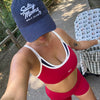 A woman takes a selfie outdoors wearing a navy blue 'Salty Moms Golf Club' baseball cap, a red sports bra with white and navy trim, and matching red athletic shorts. She is standing next to a stroller with a golf-themed pattern.