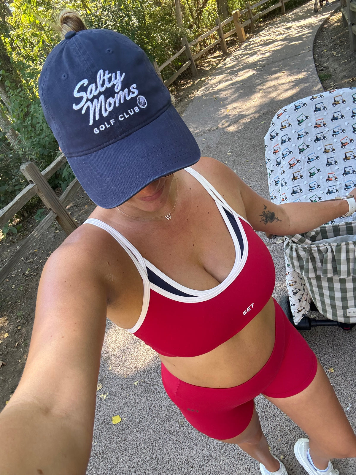 A woman takes a selfie outdoors wearing a navy blue 'Salty Moms Golf Club' baseball cap, a red sports bra with white and navy trim, and matching red athletic shorts. She is standing next to a stroller with a golf-themed pattern.