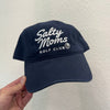 A navy blue baseball cap with white embroidered text reading 'Salty Moms Golf Club' is held by a hand against a textured off-white wall.