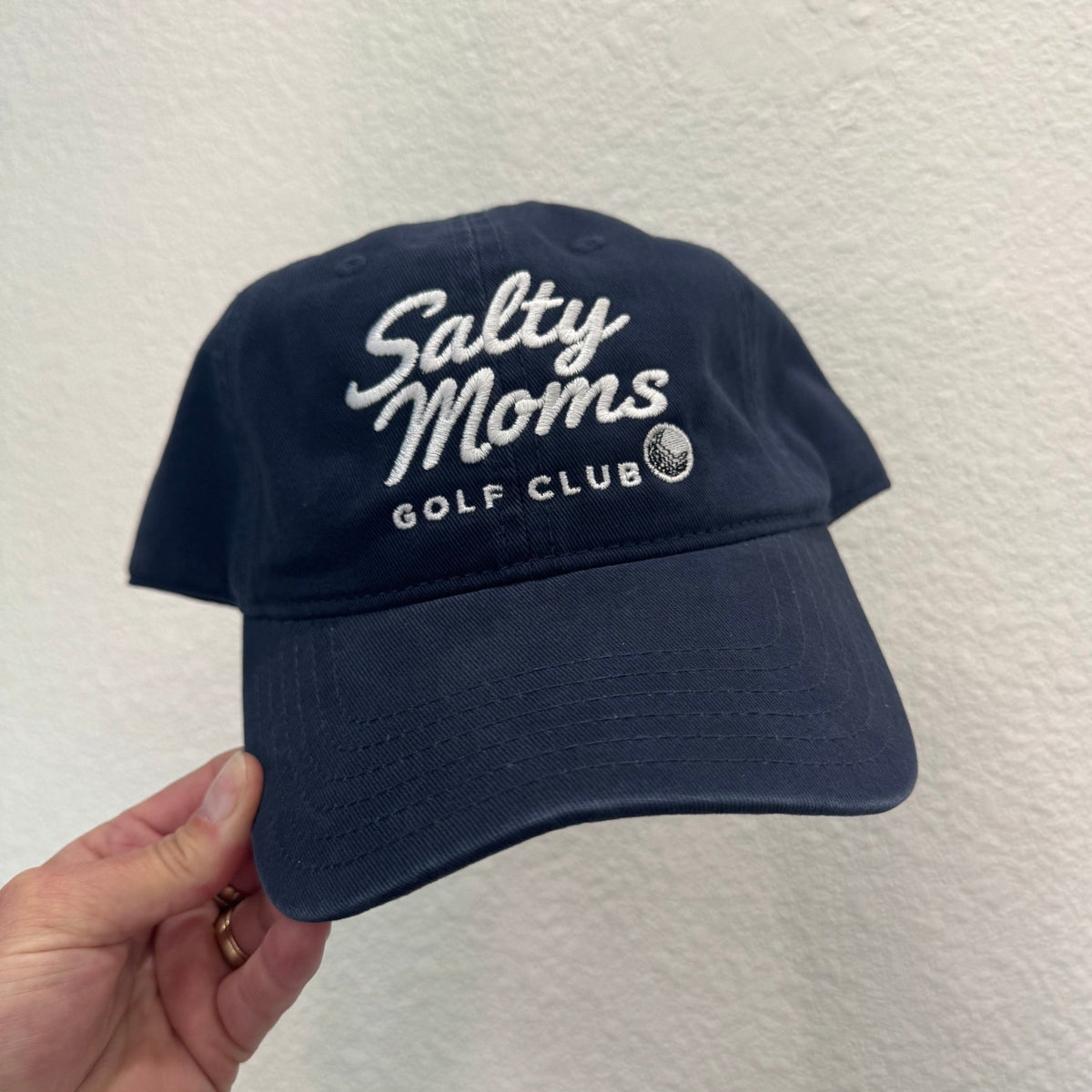 A navy blue baseball cap with white embroidered text reading 'Salty Moms Golf Club' is held by a hand against a textured off-white wall.