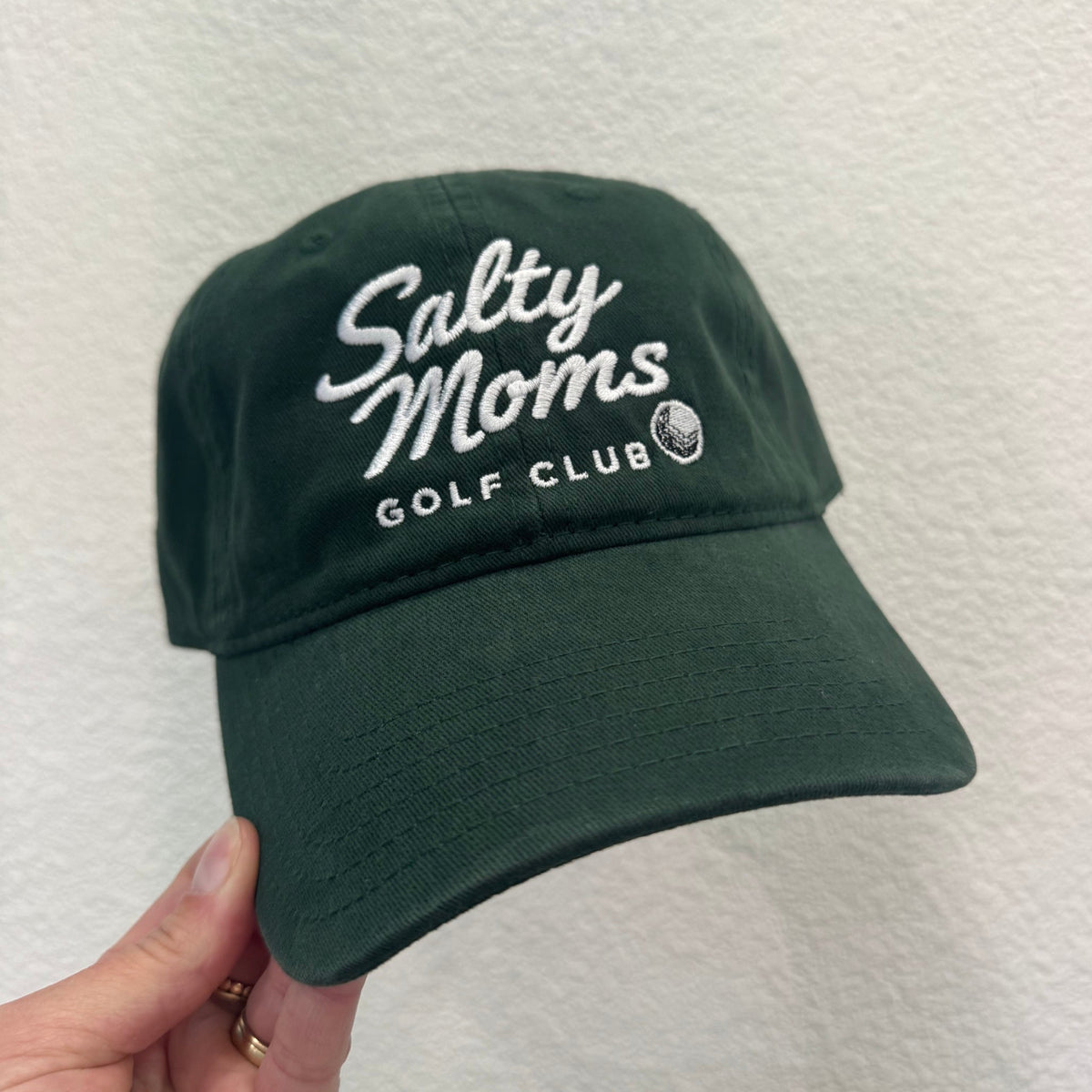 A dark green baseball cap with white embroidered text reading 'Salty Moms Golf Club' held by a hand against a textured off-white wall.