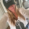 A woman taking a mirror selfie wearing a dark green baseball cap with 'Salty Moms Golf Club' embroidered in white, a cream-colored textured knit cardigan, and a dark green top. The background shows a home interior with a mirror reflecting her profile.