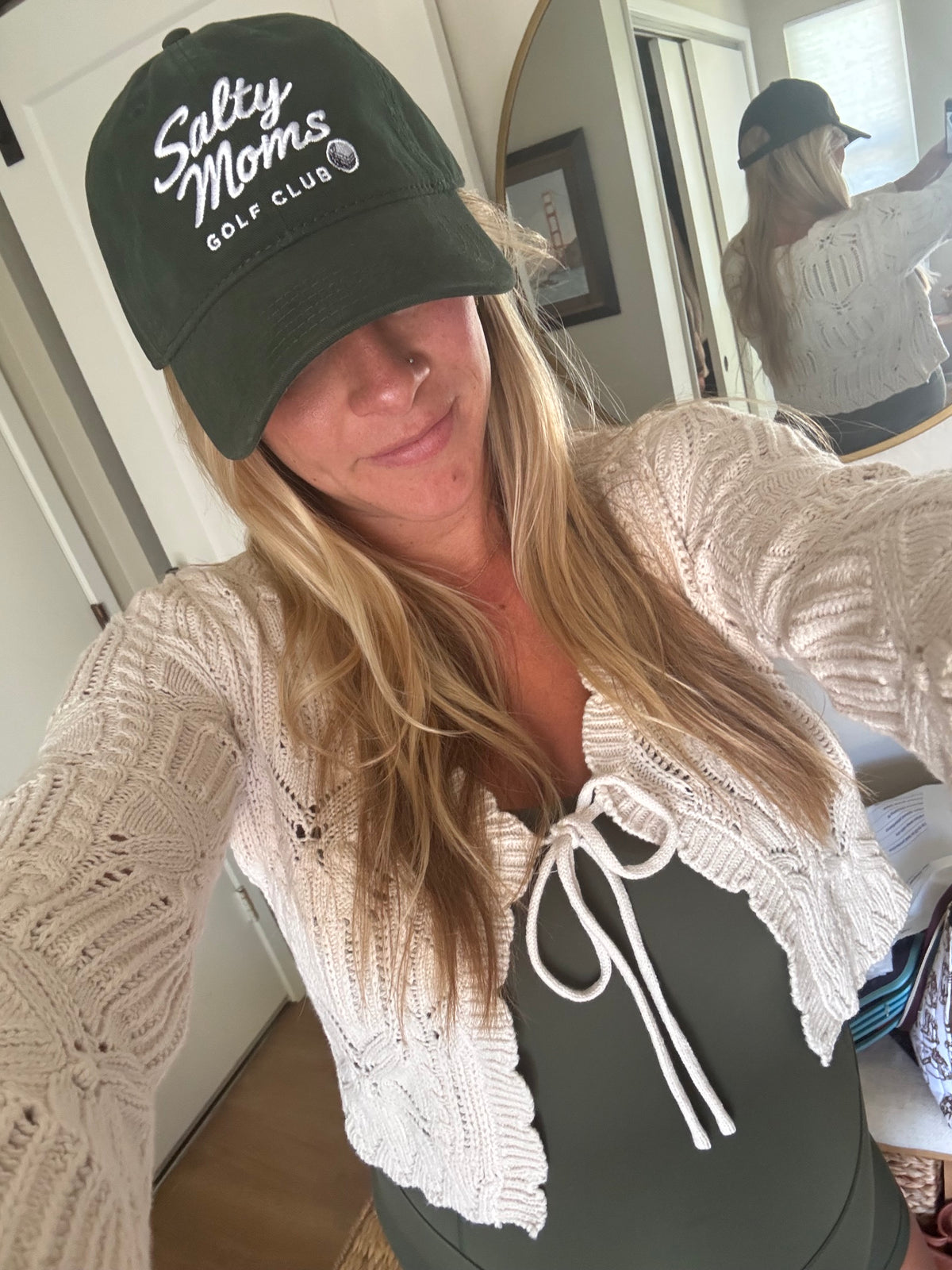 A woman taking a mirror selfie wearing a dark green baseball cap with 'Salty Moms Golf Club' embroidered in white, a cream-colored textured knit cardigan, and a dark green top. The background shows a home interior with a mirror reflecting her profile.