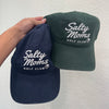 Two baseball caps held up by a hand against a textured white wall. One cap is navy blue and the other is dark green, both featuring white embroidered text that reads 'Salty Moms Golf Club'.
