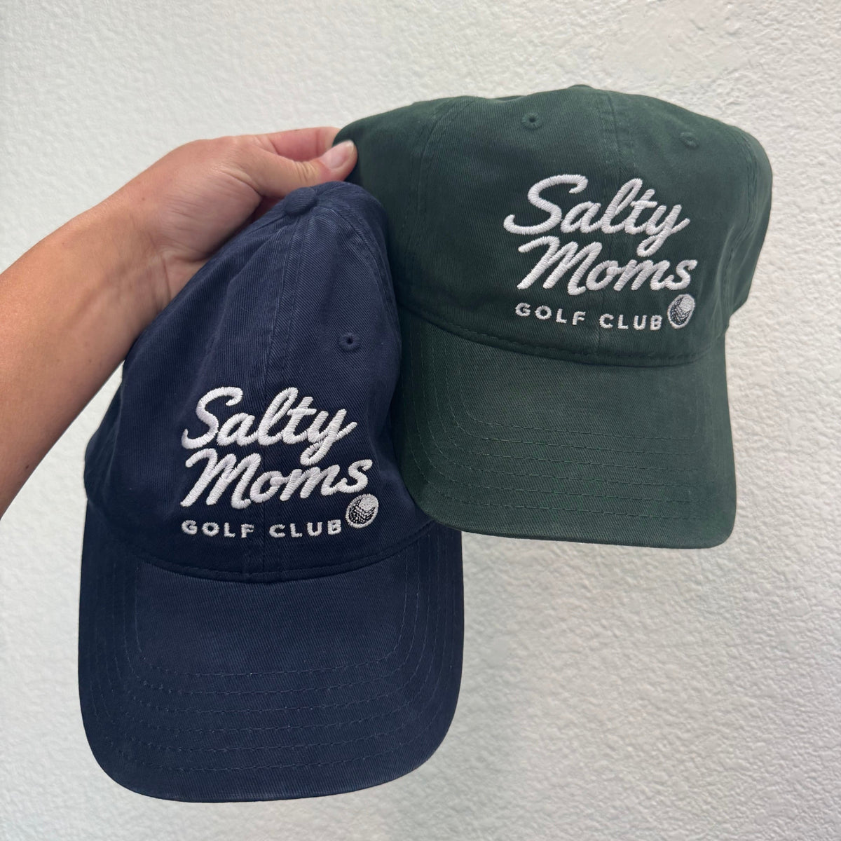 Two baseball caps held up by a hand against a textured white wall. One cap is navy blue and the other is dark green, both featuring white embroidered text that reads 'Salty Moms Golf Club'.